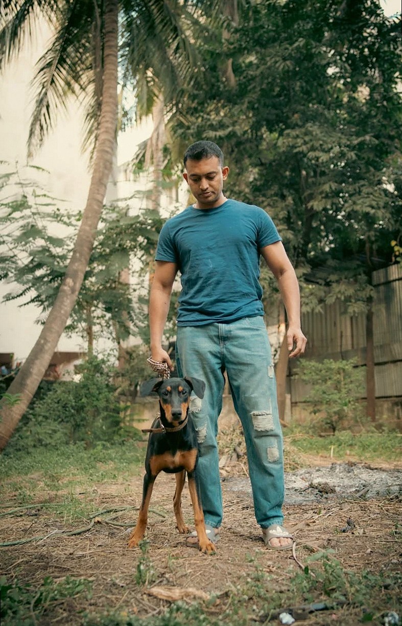 A man in a blue shirt stands outdoors with a Doberman in a natural setting.